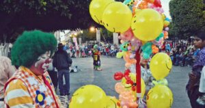 Fröhlicher Clown mit grüner Perücke und bunter Weste posiert neben lachenden Smiley-Luftballons auf einem belebten Platz beim Straßenfest; im Hintergrund unterhält ein weiterer Artist die Menge in familiärer, energiegeladener Festatmosphäre.