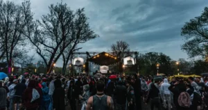 Menge bei Techno-Festival 2025 im Regen: Regenschirme, Kapuzen und Raver vor beleuchteter Bühne unter Bäumen – unerschütterliches Gemeinschaftsgefühl trotz Wetter.