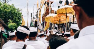 Teilnehmer einer traditionellen Straßenprozession in weißer Kleidung und Kopfbinden ziehen durch die Gassen; goldene Zeremonialschirme, Fahnen und feierliche, meditative Festivalstimmung,