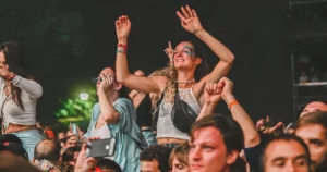 Ekstatische Menge bei Techno-Festival 2025: lachende Raver mit Glitzer-Facepaint, erhobenen Händen und Festival-Bändern – pure Energie und Gemeinschaftsgefühl in der Nacht.