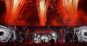 Feuerwerk über Techno-Bühne 2025: rote Explosionen, geometrische LED-Visuals und jubelnde Menge mit Leuchtstäben – episches Finale in deutscher Festkultur.