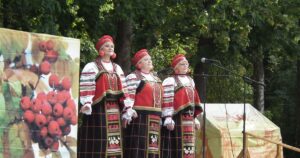 Drei Frauen in farbenfrohen ukrainischen Trachten singen leidenschaftlich auf einer Bühne vor Kalina-Beeren-Motiv, umgeben von Wald, und vermitteln die lebendige Atmosphäre eines traditionellen Folklore-Festivals in Kiew mit Gemeinschaftsgeist.
