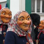 Drei lachende Holz-Hexenmasken mit Kopftuch und Strickjacke marschieren im schwäbisch-alemannischen Faschingsumzug vor weißer Hausfassade; traditionelle Narrenfiguren strahlen gruselig-fröhliche Fastnacht-Atmosphäre aus.