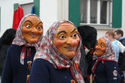 Drei lachende Holz-Hexenmasken mit Kopftuch und Strickjacke marschieren im schwäbisch-alemannischen Faschingsumzug vor weißer Hausfassade; traditionelle Narrenfiguren strahlen gruselig-fröhliche Fastnacht-Atmosphäre aus.