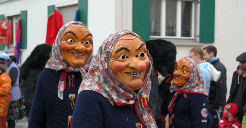 Drei lachende Holz-Hexenmasken mit Kopftuch und Strickjacke marschieren im schwäbisch-alemannischen Faschingsumzug vor weißer Hausfassade; traditionelle Narrenfiguren strahlen gruselig-fröhliche Fastnacht-Atmosphäre aus.